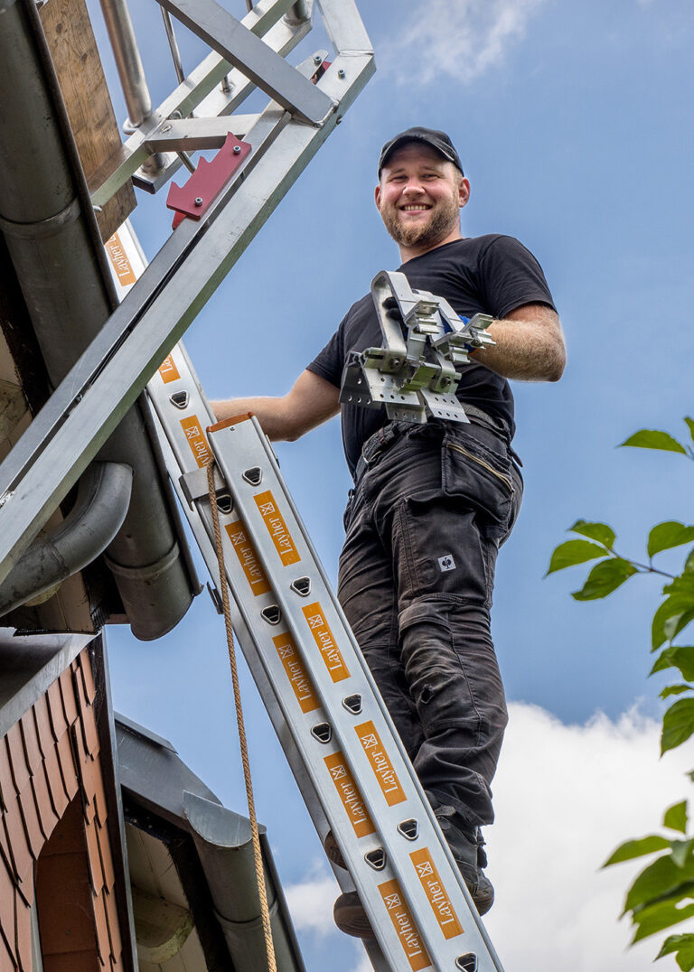 Ein lächelnder Bauarbeiter klettert eine Leiter hinauf und trägt Metallhalterungen, vor einem klaren blauen Himmel.