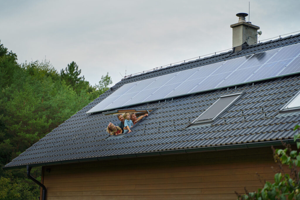 Eine glückliche Familie schaut aus einem Dachfenster eines Hauses mit eine Solaranlage auf dem Dach.